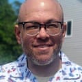 A person with a salt-and-pepper beard who is wearing glasses poses outside