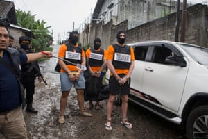 three people in orange wear handcuffs on a muddy street