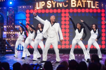 In a white dinner jacket, he points dramatically in the air; five backing dancers copy him