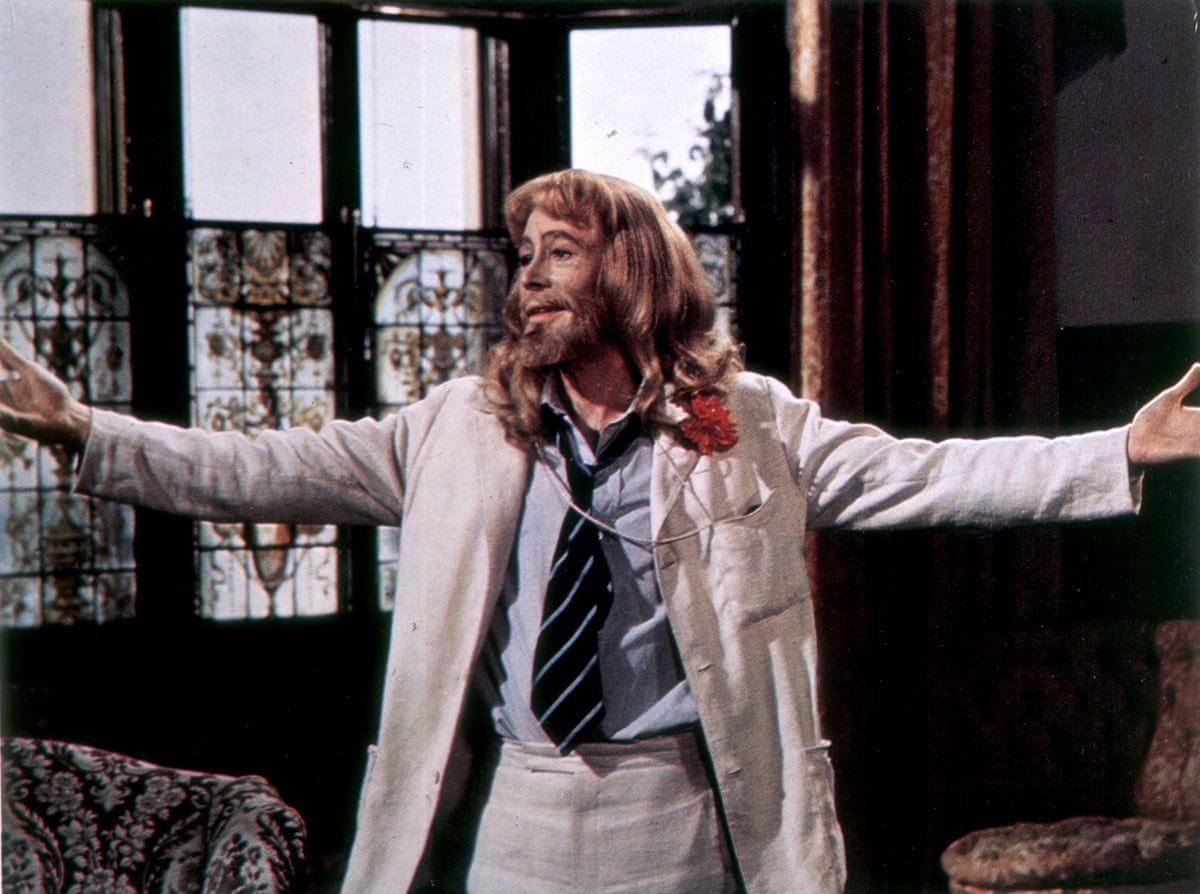 Peter O'Toole, wig-bearded and in a cream suit, stands smiling with arms outstretched