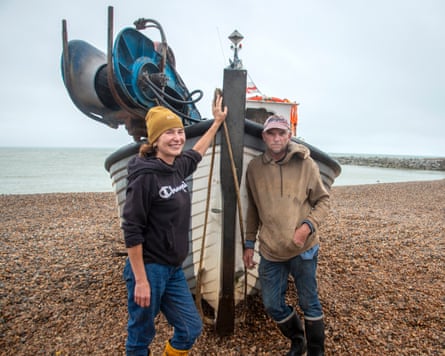 A man and a woman stand either side of the prow of a wooden fishing boat on a shingle beach