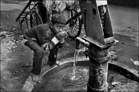 A boy crouches down as he scoops water into his mouth from a hand pump