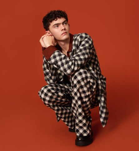 Joe Locke, with short dark curly hair, wearing a black-and-white check suit, a dark red shirt and shiny black shoes, and crouching in front of a dark orange background