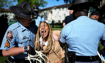 A young Black woman with long brown and white braids screams as two police officers in blue shirts and black hats hold her arms behind her.