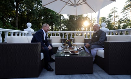 Narendra Modi y Vladimir Putin se sientan afuera en una terraza con columnas blancas en la residencia oficial de Putin; están sentados en sofás bajos con una mesa de café entre ellos y una sombrilla blanca sobre sus cabezas; hay árboles detrás de ellos y el sol brilla a través de las ramas.