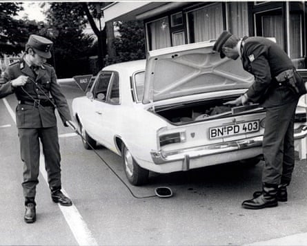 Two men, one looks in a car boot, the other uses a mirror to look under car itself