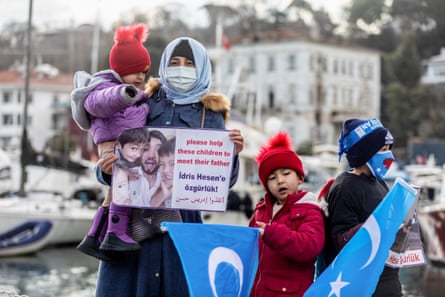 A woman and three children outside. Buildings and boats can be seen behind them. She holds a sign with a photograph of a man and three children that reads ‘please help these children to meet their father’