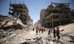 people walk amid concrete rubble and dust beside multistory buildings with blown-out walls, under clear blue sky.