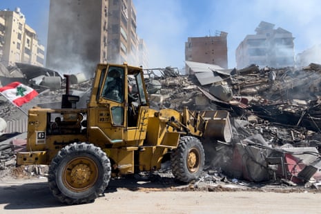 A yellow tractor drives in front of the debris of a building, dust rising into the blue sky.