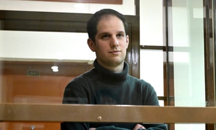 Evan Gershkovich, wearing a black turtleneck, stands in a glass cage in a courtroom at the Moscow City Court with his arms crossed. 
