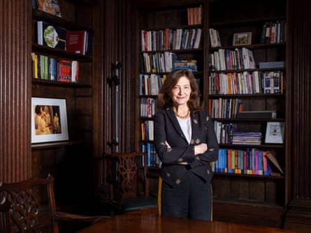 Prof Deborah Prentice stands in a dark study with shelves of books behind her. She appears to be in her 60s with wavy dark brown hair; she wears a white blouse, black trousers and a short black jacket with a gold leaf pattern on it, plus a chunky gold chain necklace.