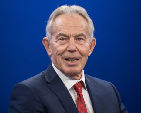Tony Blair in a dark suit and red patterned tie smiles in front of a blue backdrop