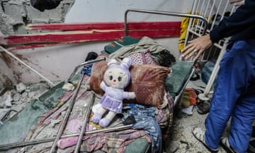 A medic almost off-camera gestures at a child's soft toy lying on a hospital bed amid rubble and wreckage from an airstrike