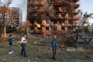 Three people stand in front of a burnt out, smoking building. Parts of the building are still on fire
