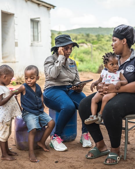 Two women sit on chairs outside a small house. One has a small child on her lap. There are two other small children next to them