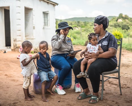 Two women sit on chairs outside a small house. One has a small child on her lap. There are two other small children next to them