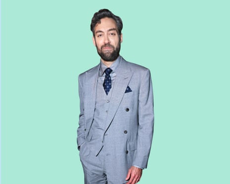 Alex Hassell wearing a grey three-piece suit standing with his right hand in his pocket against a duck-egg background