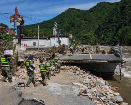 Soldiers in hi-vis vests carry boxes across a broken section of road beside a river