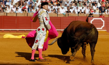 A matador in a white costume holding a bright pink cape next to a bull in motion in a bullring.