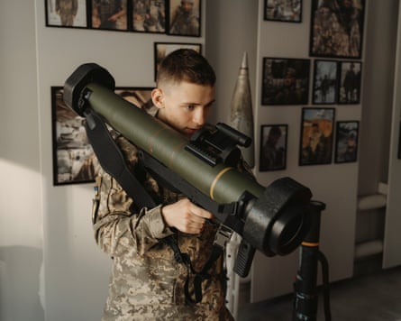 A cadet holds an anti-tank missile launcher, a large cylindrical black weapon, across his shoulder and looks down its sights.