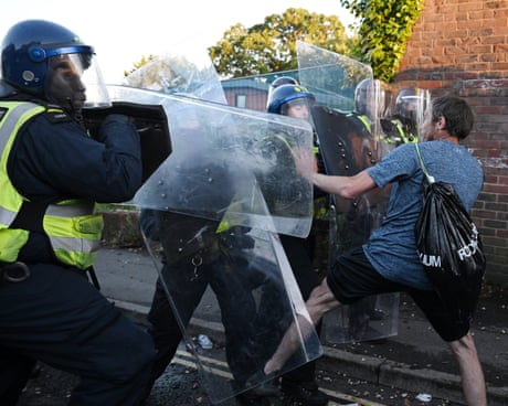 Riot police with shields confront a man on a street near a brick wall.