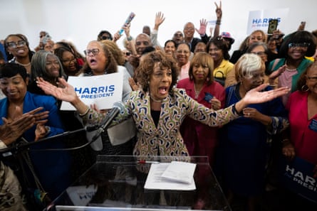 Older Black woman in patterned vest gestures outward with both hands at a lectern in front of a crowd of jubilant-appearing Black women.