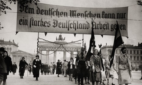 A column of people, led by two women carrying Nazi flags, and others  a huge banner saying 'Dem deutschen Menschen kann nur ein starkes Deutschland Arbeit geben'