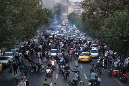 A wide street crowded with motorbikes and cars with a fire in the background 