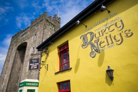 A bright yellow pub building, a green and cream phone box and a tall stone castle gateway against a blue sky