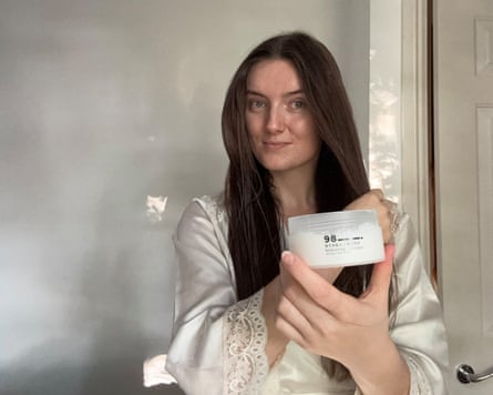 The writer holds up a pot of hair mask while her other hand is touching her hair