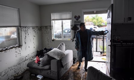 a man in a poncho and boots stands in a home where there is debris from flooding and a line of dirt on the walls from the height of the water