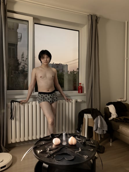 Photograph of Enkiny wearing only pants, leaning against a radiator with an open window behind him. On the table in front of him are his sex toys.