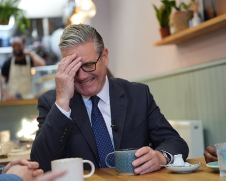 Starmer laughs with his hand to his forehead while sitting at a cafe table