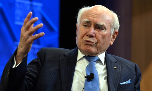 A close-up view of John Howard as he speaks at the National Press Club. His lips are pursed and he is gesturing with one hand. There is a microphone attached to his blue tie and he is wearing a dark blue suit. The press club logo can be seen on a screen behind him