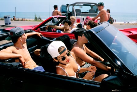 A young woman in backwards baseball cap, bikini and sunglasses in an open-topped car with two young men , the sea visible behind