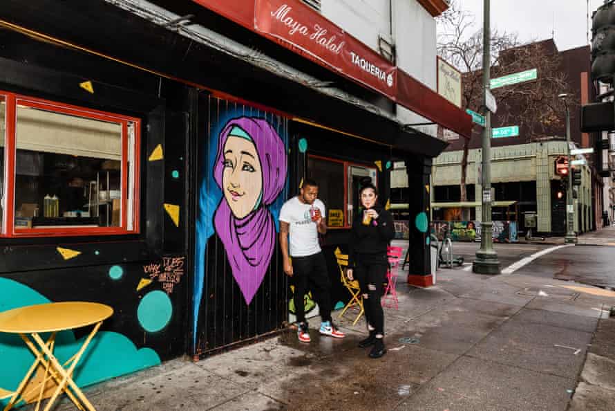 A man and woman sip on a drink outside a restaurant with a mural of a woman in a hijab