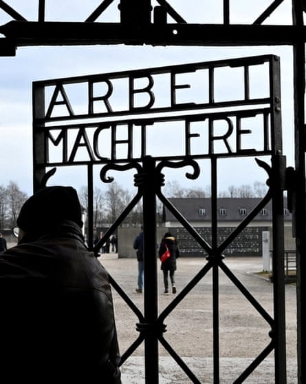 A visitor to Dachau stands at the main gate that says Arbeit Macht Frei