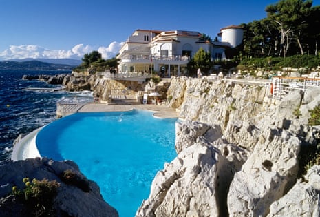 View across blue infinity pool on a cliff edge towards a white hotel building with the sea to one side.