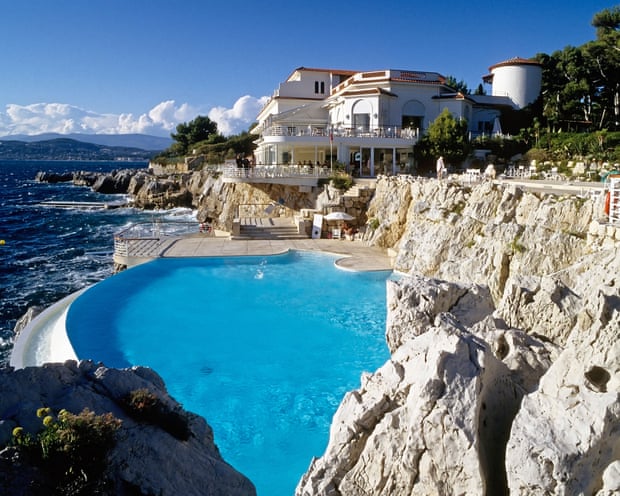 View across blue infinity pool on a cliff edge towards a white hotel building with the sea to one side.