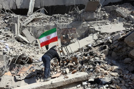 A man dressed in a black shirt and blue jeans faces away from the camera, sticking the pole of the Iranian flag atop the ruins of a building.