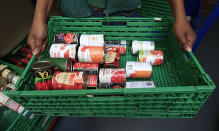 A food bank worker holds a green plastic container that holds various tins of plum and chopped tomatoes