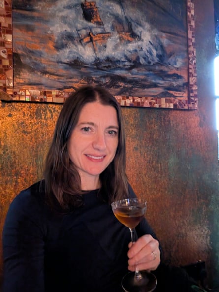 A woman with long dark hair smiling and holding a drink in a tall-stemmed glass with a picture of a boat in a storm on the wall behind her