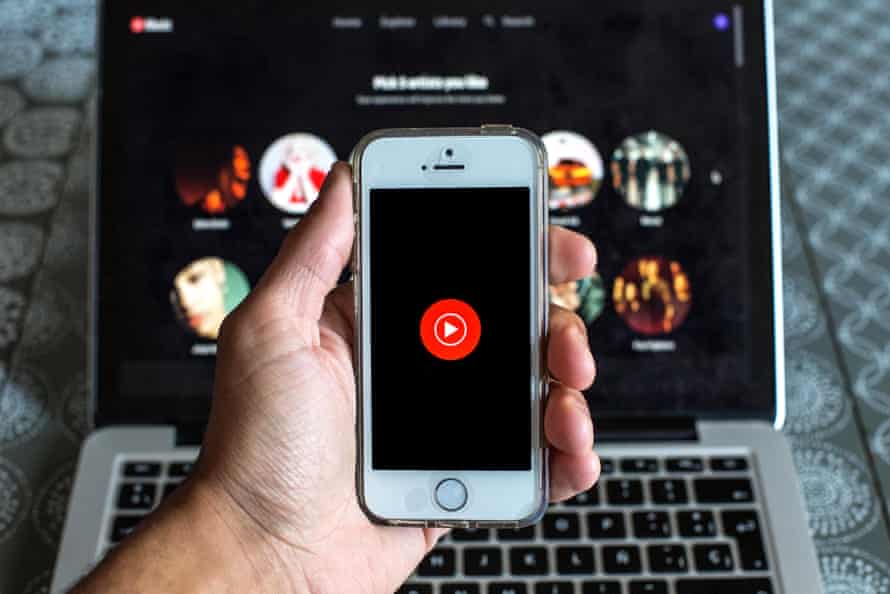 Hand holding a phone in front of a laptop, with both displaying YouTube Music apps