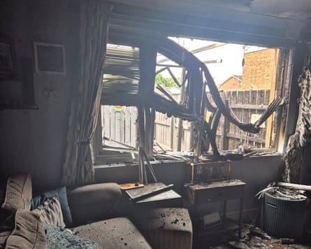 The burned-out living room and broken back window; there is debris over the blackened grey sofa, the floor and other furniture.