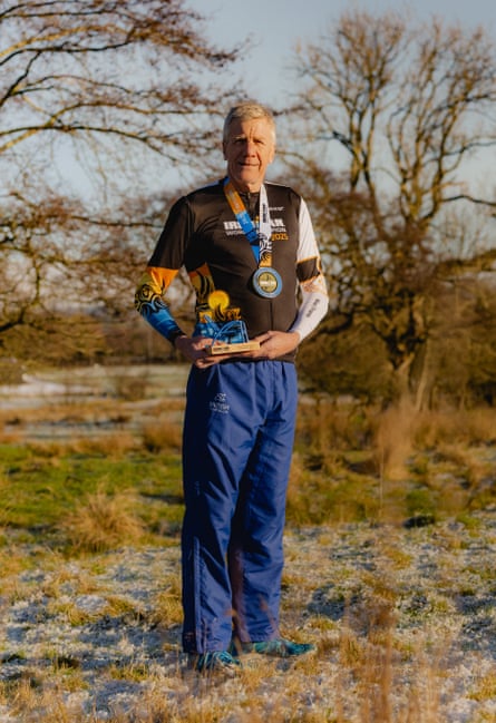 A man wearing dark-blue tracksuit bottoms, a sporty brown top and blue trainers, standing on some shrubland wearing a medal and holding a trophy