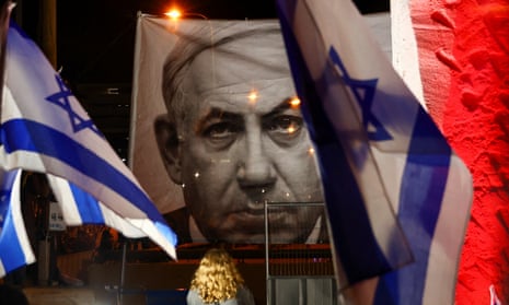 A person stands in front of a large banner depicting Benjamin Netanyahu during a protest in Tel Aviv