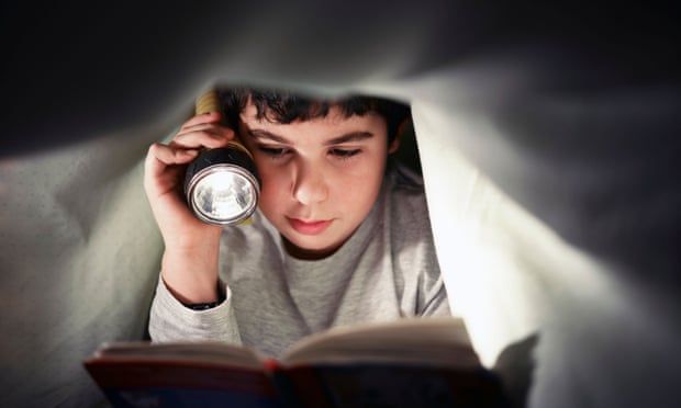 A boy reading a book under the bed covers.