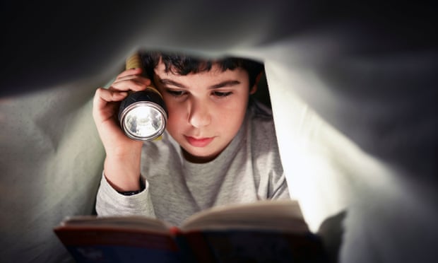 A boy reads a book under bed covers using a torch