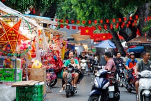 A woman and a child ride through a street market with brightly coloured stalls and red bunting featuring the Vietnamese flag.