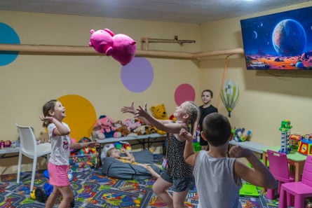 Two girls laugh as they throw a pink soft toy to each other in a play room as two boys look on, while another girl lies on a bean bag